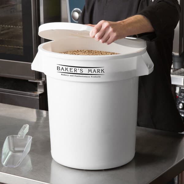 A woman pouring grains into a large white Baker's Mark ingredient storage container with a lid.