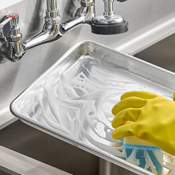 A small aluminum bun or sheet pan being hand-washed in a sink.