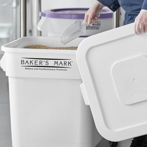 A person using a plastic scoop to fill a Baker's Mark white ingredient storage bin with a lid.