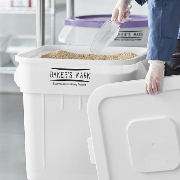 A person using a gloved hand to put food in a white Baker's Mark ingredient storage bin.