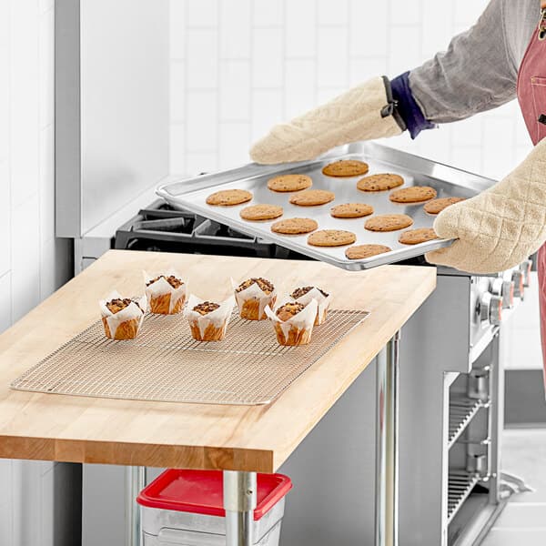 A woman in an apron holding a Baker's Mark wire-in-rim aluminum sheet pan with cookies on a counter.