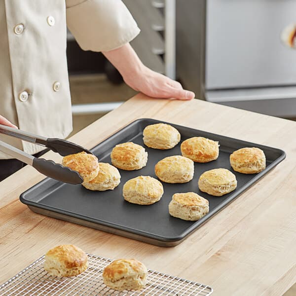 A person using tongs to take biscuits off a Baker's Mark non-stick sheet pan.