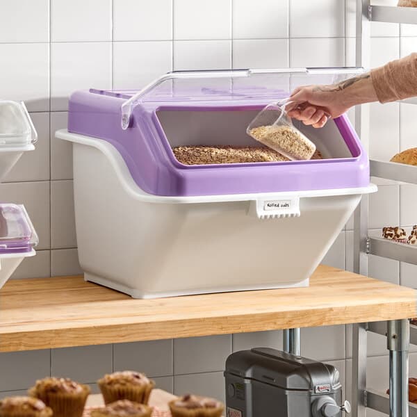 A large plastic ingredient bin with a purple flip lid, label sheet, and scoop, designed for allergen-free storage on a kitchen shelf.