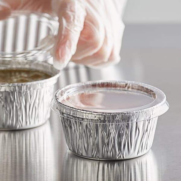 A person in a plastic glove holding a Baker's Lane round aluminum container with a plastic lid.