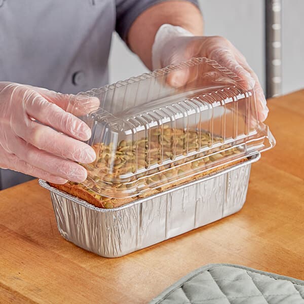 A person in gloves holding a Baker's Lane aluminum foil loaf pan filled with food.