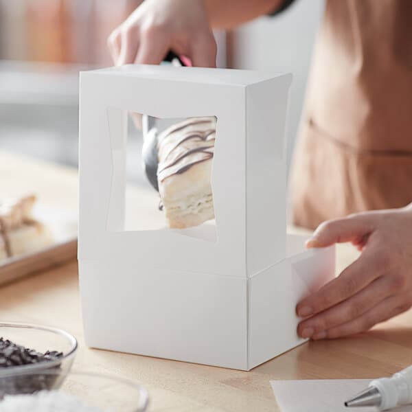 A white bakery box with a clear window on the side, being used to package a dessert.