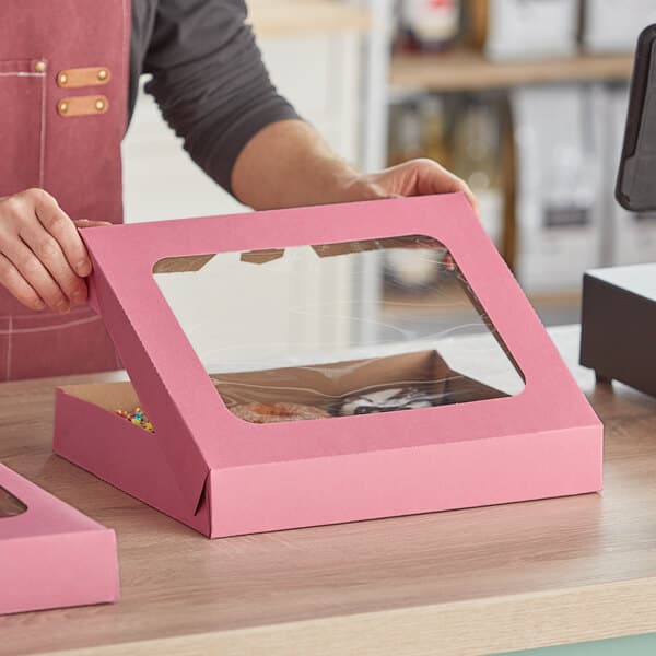 A woman holding a pink Baker's Mark donut box with a clear window.