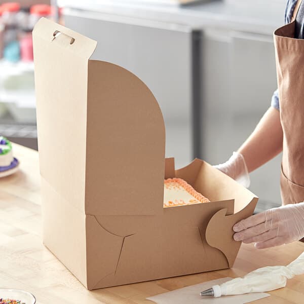 A woman opening a Baker's Mark kraft cake box on a counter to reveal a cake with orange frosting.