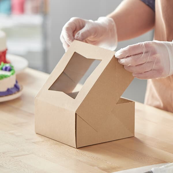 A kraft-colored bakery box with a clear window on the lid being assembled on a wooden surface.