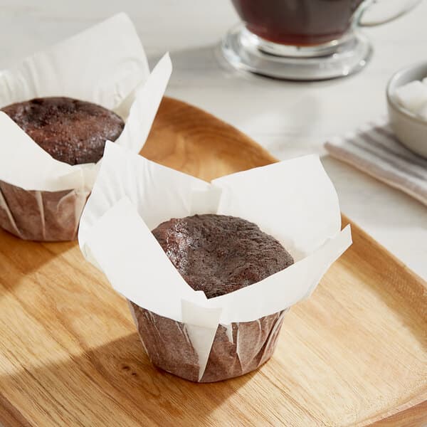 A chocolate muffin in a white high crown muffin wrap on a wooden tray.