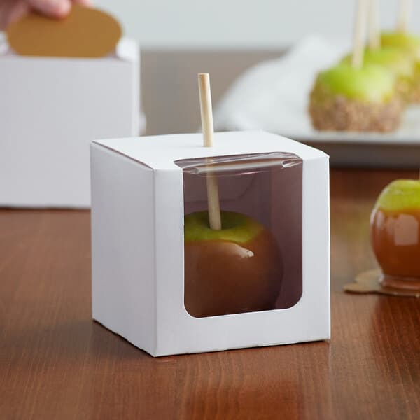 A white Baker's Lane candy apple box with a window holding a green apple with a wooden stick.