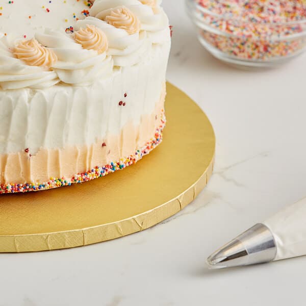 A cake with frosting and sprinkles on a 12" gold cake drum on a table with a white bag with a silver tip.