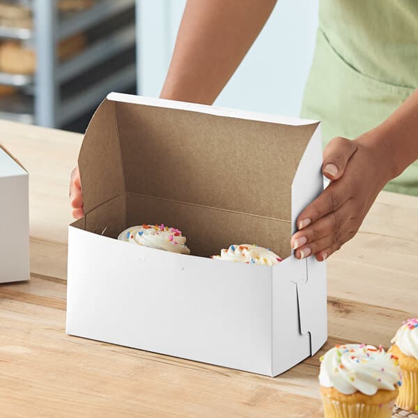 A white bakery box partially filled with cupcakes, being assembled on a wooden surface.