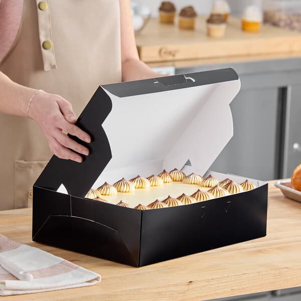 A black quarter sheet cake bakery box with a cake inside, being opened on a kitchen counter.