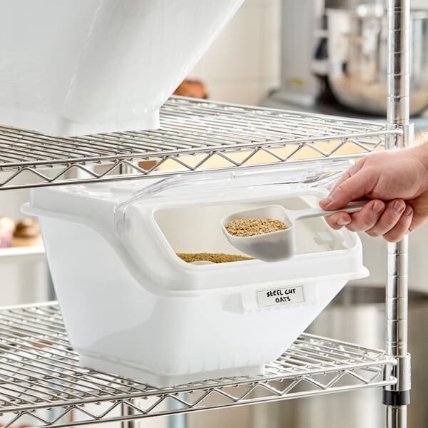 A white plastic ingredient bin with a flip lid, label, and scoop, used for storing bulk dry goods on a wire shelf.